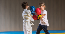Two children in karate attire practicing boxing with gloves indoors.