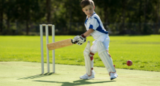 A young boy in cricket gear playing a shot on a sunny field.