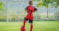 Child in a red soccer kit with number 7 standing with a ball on a green field facing goalposts.