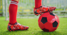 Soccer player with red cleats resting foot on a black and red soccer ball.