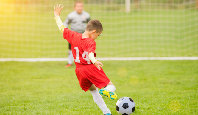 Child in red jersey kicking soccer ball on field.